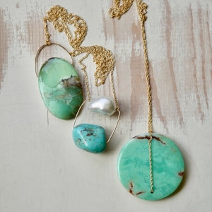 Puede incluir: Un collar de cadena dorada con cuatro colgantes de piedras preciosas verdes y azules. El collar presenta una piedra verde redonda grande, una piedra verde ovalada m&aacute;s peque&ntilde;a, una piedra turquesa y una perla blanca.