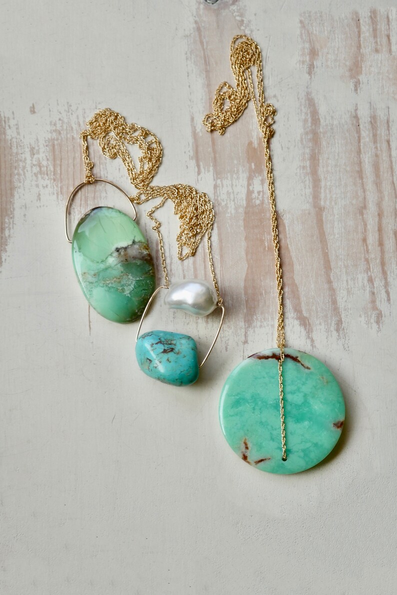 Puede incluir: Un collar de cadena dorada con cuatro colgantes de piedras preciosas verdes y azules. El collar presenta una piedra verde redonda grande, una piedra verde ovalada m&aacute;s peque&ntilde;a, una piedra turquesa y una perla blanca.