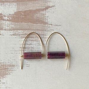May include: A pair of gold hoop earrings with a rectangular purple stone accent. The earrings are simple and elegant.