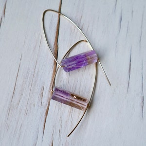 May include: A pair of dangle earrings featuring elongated, faceted purple and tan crystals suspended from delicate gold-colored wires. The earrings are displayed against a white, weathered wooden surface, creating a contrast of textures and colors.