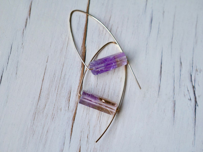 May include: A pair of dangle earrings featuring elongated, faceted purple and tan crystals suspended from delicate gold-colored wires. The earrings are displayed against a white, weathered wooden surface, creating a contrast of textures and colors.