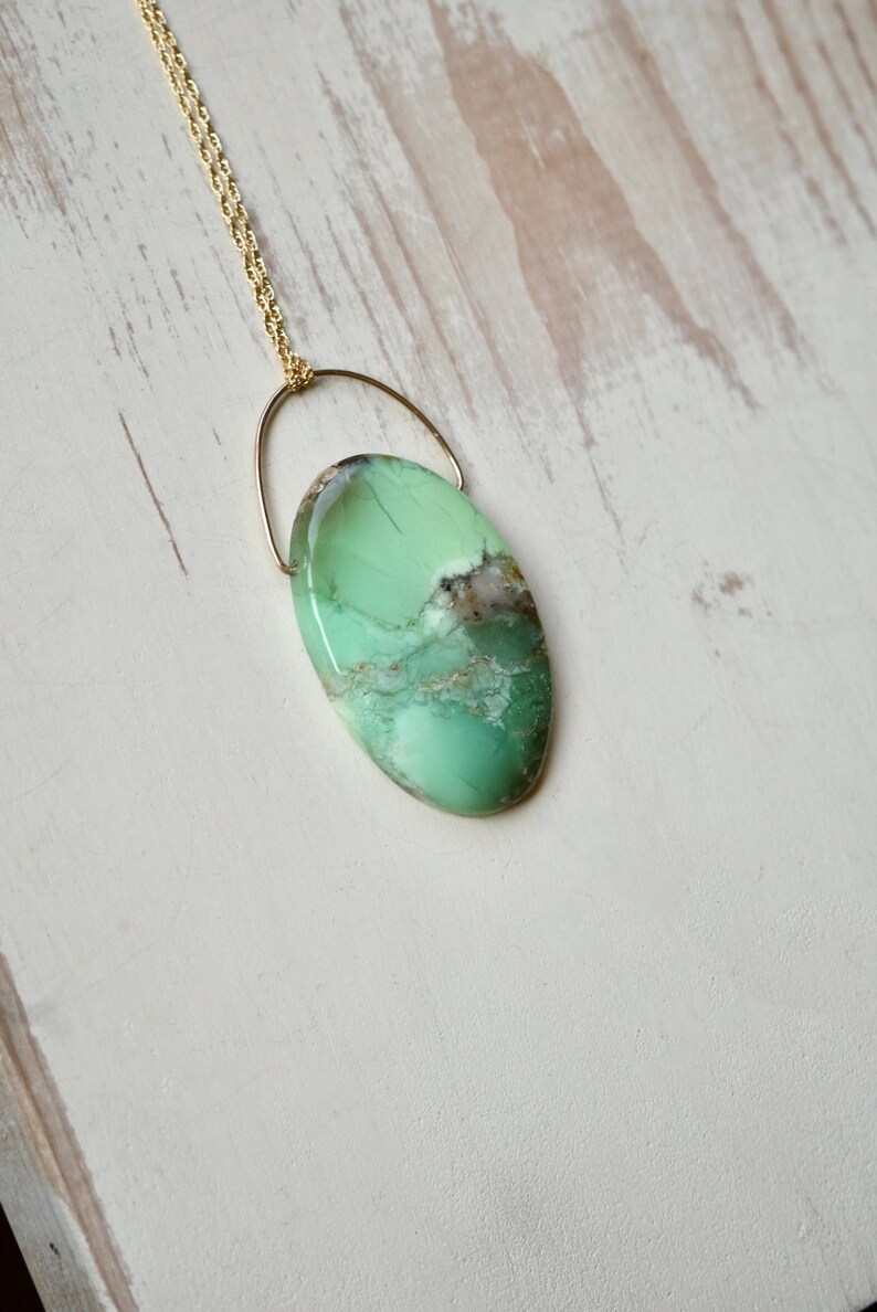 Puede incluir: Un collar de cadena dorada con un colgante ovalado verde. El colgante est&aacute; hecho de una piedra verde con vetas blancas y marrones.