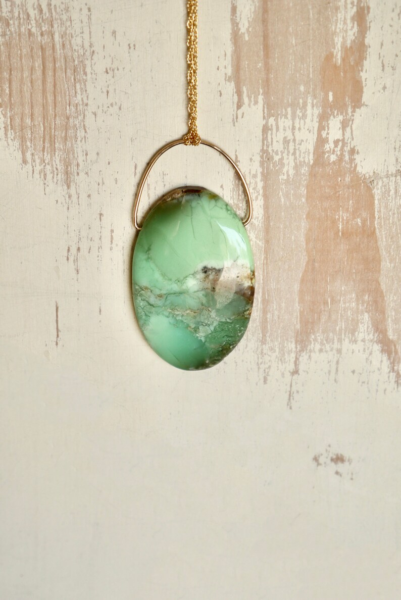 Puede incluir: Un collar de cadena dorada con un colgante ovalado verde y marr&oacute;n. El colgante est&aacute; engastado en un marco de alambre dorado.