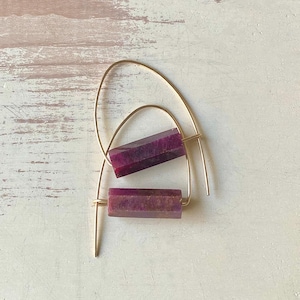 May include: A pair of gold hoop earrings with a purple gemstone bar pendant. The earrings are simple and elegant.
