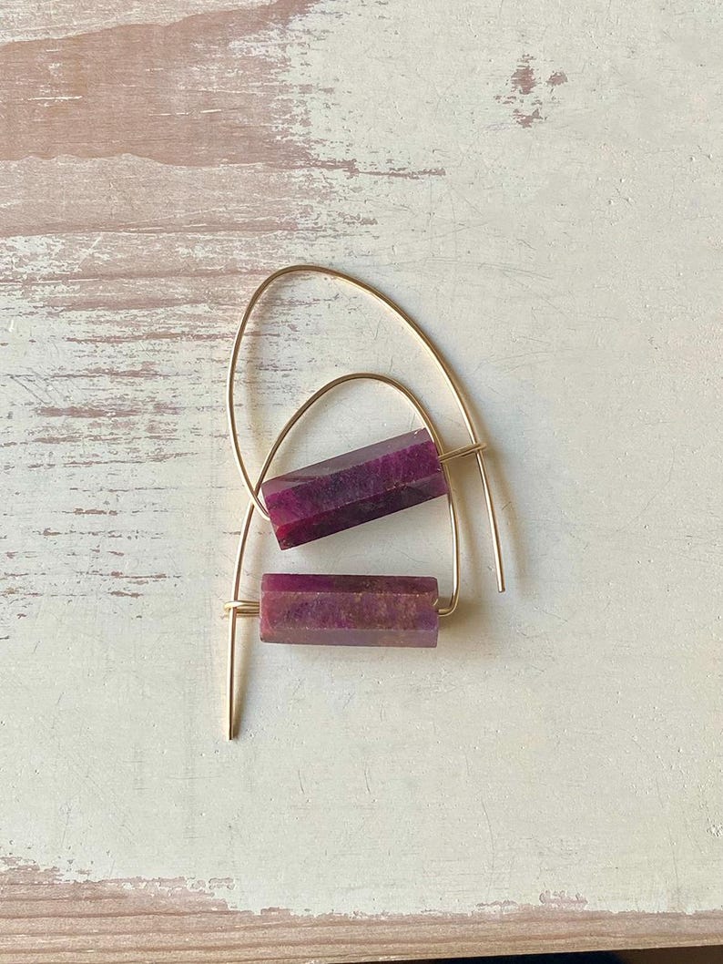 May include: A pair of gold hoop earrings with a purple gemstone bar pendant. The earrings are simple and elegant.