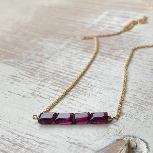 May include: A gold chain necklace with a bar pendant made of five square-shaped, dark purple gemstones.