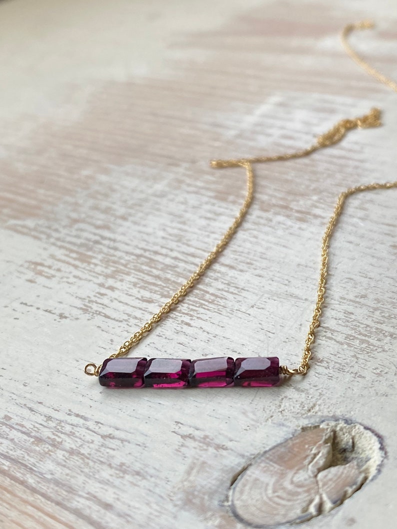 May include: A gold chain necklace with a bar pendant made of five square-shaped, dark purple gemstones.