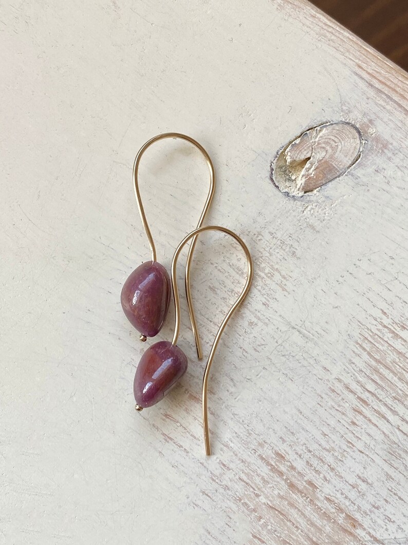 May include: A pair of gold dangle earrings with a deep purple gemstone. The earrings are simple and elegant.