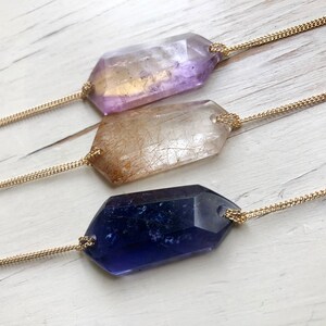 May include: Three gold chain necklaces with different colored crystal pendants. The pendants are all faceted and have a geometric shape. The crystals are purple, clear, and blue.