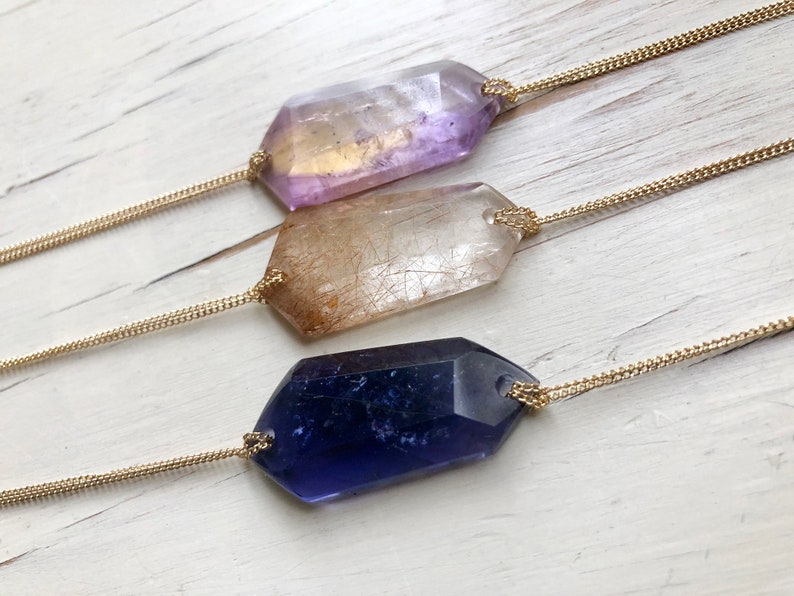 May include: Three gold chain necklaces with different colored crystal pendants. The pendants are all faceted and have a geometric shape. The crystals are purple, clear, and blue.