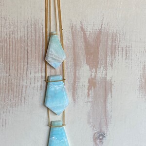 Pu&ograve; includere: Una collana con tre pendenti in pietra preziosa azzurra e bianca. Ogni pendente ha una forma geometrica unica, sospesa a una delicata catena dorata. La collana &egrave; appesa su uno sfondo chiaro e strutturato.