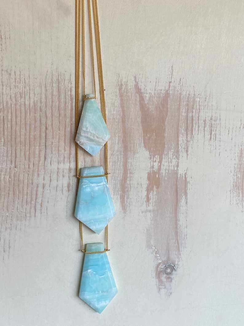 Pu&ograve; includere: Una collana con tre pendenti in pietra preziosa azzurra e bianca. Ogni pendente ha una forma geometrica unica, sospesa a una delicata catena dorata. La collana &egrave; appesa su uno sfondo chiaro e strutturato.