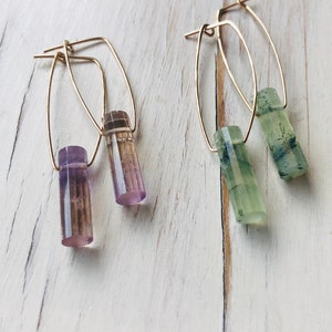 May include: A pair of gold hoop earrings with a purple and green crystal dangle. The earrings are on a white wooden surface.