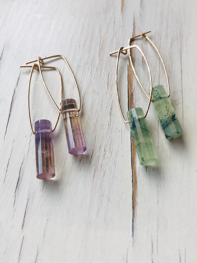 May include: A pair of gold hoop earrings with a purple and green crystal dangle. The earrings are on a white wooden surface.