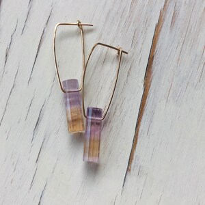 May include: A pair of gold hoop earrings with a purple and yellow gemstone dangle. The earrings are on a white wooden surface.