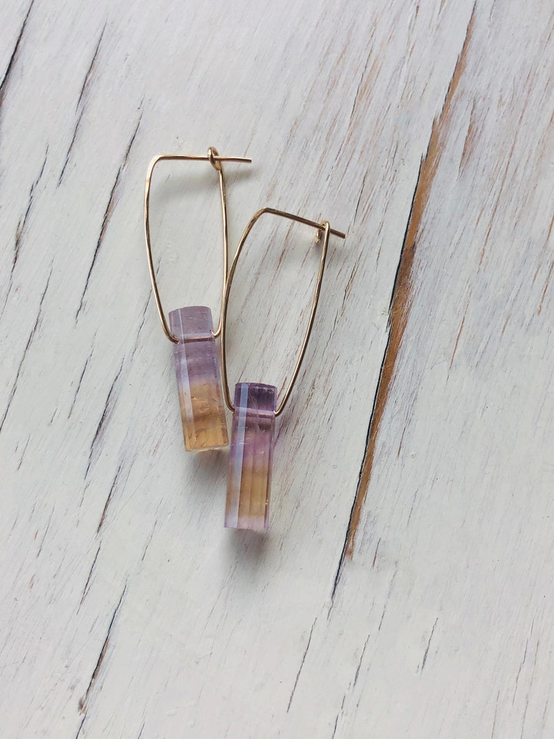May include: A pair of gold hoop earrings with a purple and yellow gemstone dangle. The earrings are on a white wooden surface.