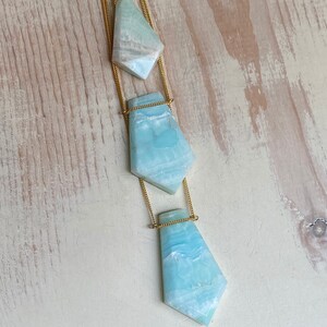 Pu&ograve; includere: Una collana con catena dorata con tre pendenti in pietra azzurra di forma irregolare. I pendenti sono collegati da sottili catene dorate, creando un effetto a strati. Le pietre hanno un aspetto marmorizzato con sfumature bianche e blu.