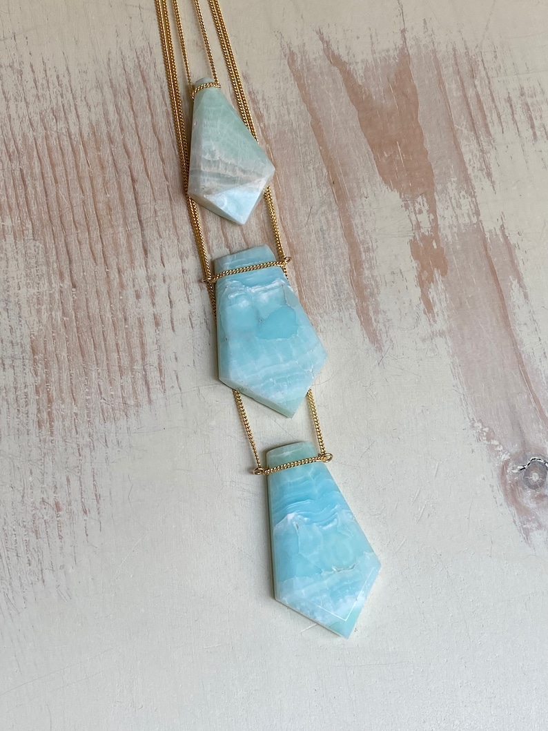 Pu&ograve; includere: Una collana con catena dorata con tre pendenti in pietra azzurra di forma irregolare. I pendenti sono collegati da sottili catene dorate, creando un effetto a strati. Le pietre hanno un aspetto marmorizzato con sfumature bianche e blu.