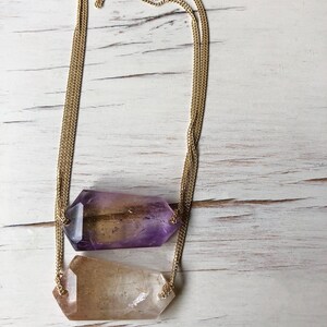 May include: A gold chain necklace with two crystal pendants. The top pendant is a light purple and the bottom pendant is a clear quartz.