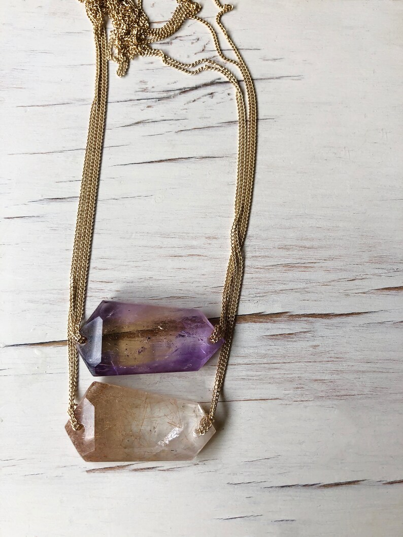 May include: A gold chain necklace with two crystal pendants. The top pendant is a light purple and the bottom pendant is a clear quartz.