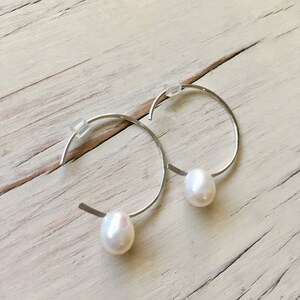 May include: A pair of silver hoop earrings with a single white pearl dangling from each hoop.