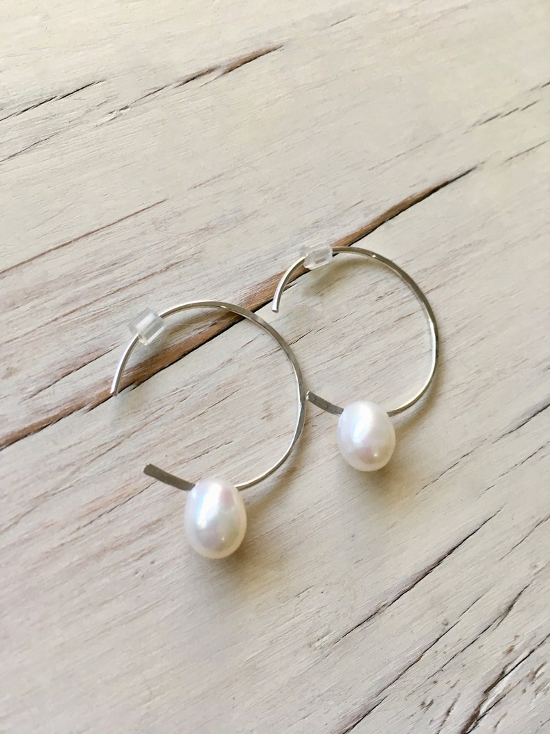 May include: A pair of silver hoop earrings with a single white pearl dangling from each hoop.