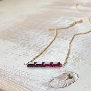 May include: A delicate gold chain necklace with a bar pendant featuring five small, square-shaped, deep purple gemstones.