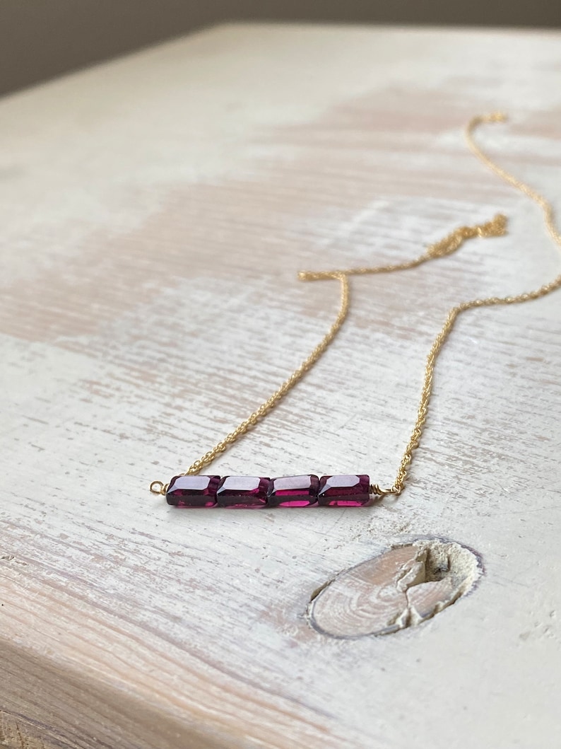 May include: A delicate gold chain necklace with a bar pendant featuring five small, square-shaped, deep purple gemstones.