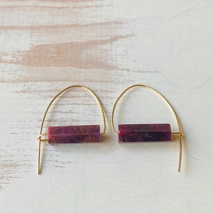 May include: A pair of gold hoop earrings with a rectangular purple stone pendant. The earrings are simple and elegant.