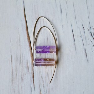 May include: A pair of gold-colored wire earrings with two rectangular, translucent, purple and brown crystal beads. The earrings are displayed on a white, distressed wooden surface.