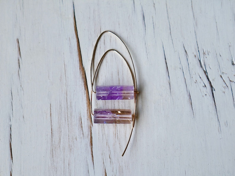 May include: A pair of gold-colored wire earrings with two rectangular, translucent, purple and brown crystal beads. The earrings are displayed on a white, distressed wooden surface.