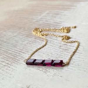May include: A delicate gold chain necklace with a bar of five square-shaped, deep purple gemstones.