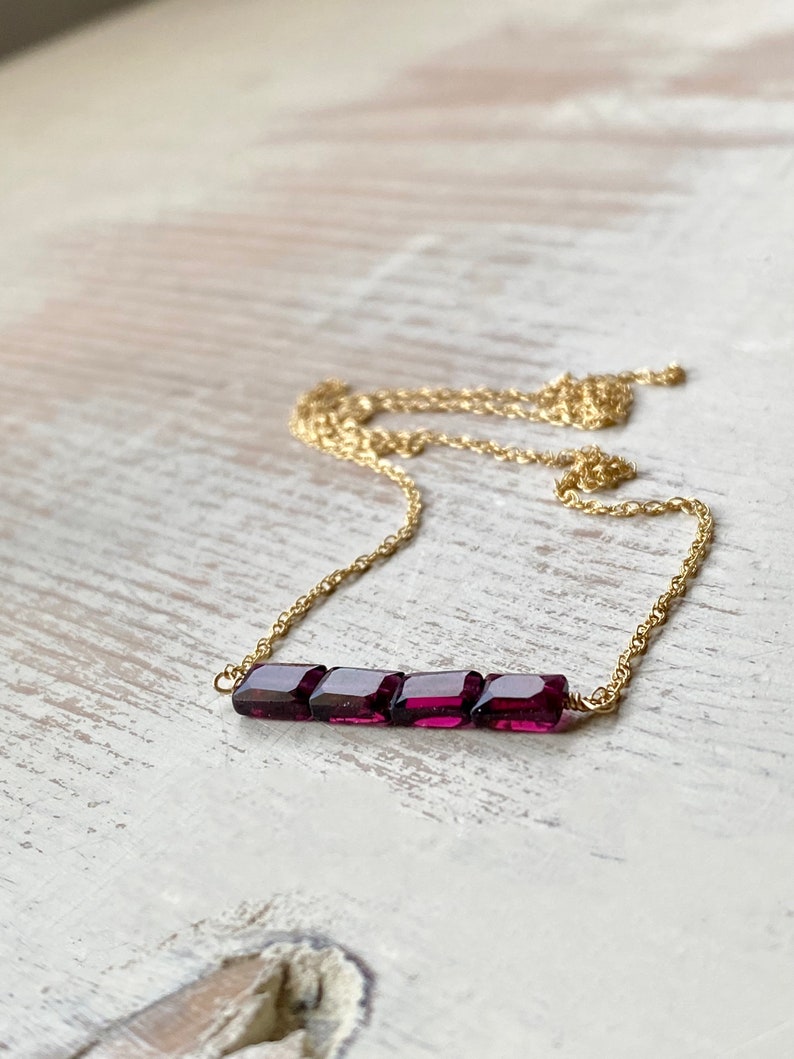 May include: A delicate gold chain necklace with a bar of five square-shaped, deep purple gemstones.