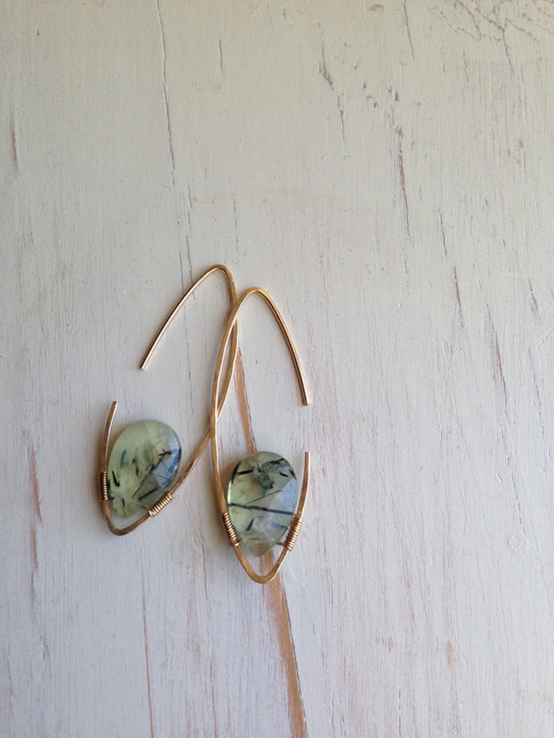 May include: A pair of gold earrings with green and black gemstone drops. The earrings are made with thin wire and have a simple, elegant design.