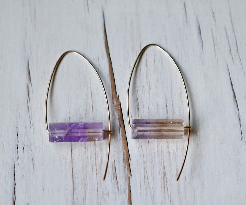 May include: A pair of gold-colored hoop earrings, each featuring a rectangular, translucent amethyst stone. The amethyst stones display shades of purple and white. The earrings are displayed on a white, textured surface.