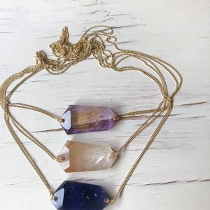 May include: A gold chain necklace with three different colored crystal pendants. The crystals are in the shape of a long, thin rectangle. The crystals are purple, clear, and blue.