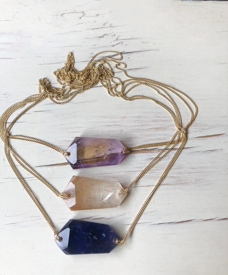 May include: A gold chain necklace with three different colored crystal pendants. The crystals are in the shape of a long, thin rectangle. The crystals are purple, clear, and blue.