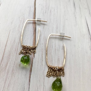 May include: Gold hoop earrings with a dangling green peridot stone and a chain fringe.