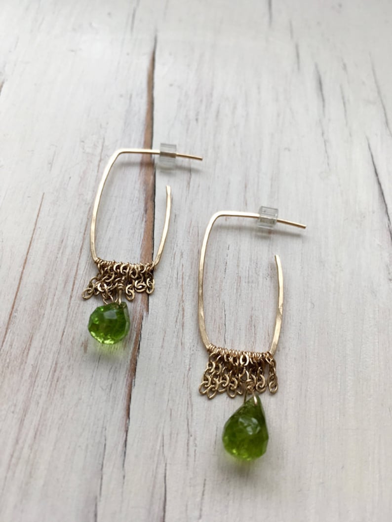 May include: Gold hoop earrings with a dangling green peridot stone and a chain fringe.