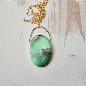 Puede incluir: Un colgante de piedra preciosa ovalado de color verde y marr&oacute;n en una cadena de oro. El colgante est&aacute; suspendido de un fino bucle de alambre de oro.