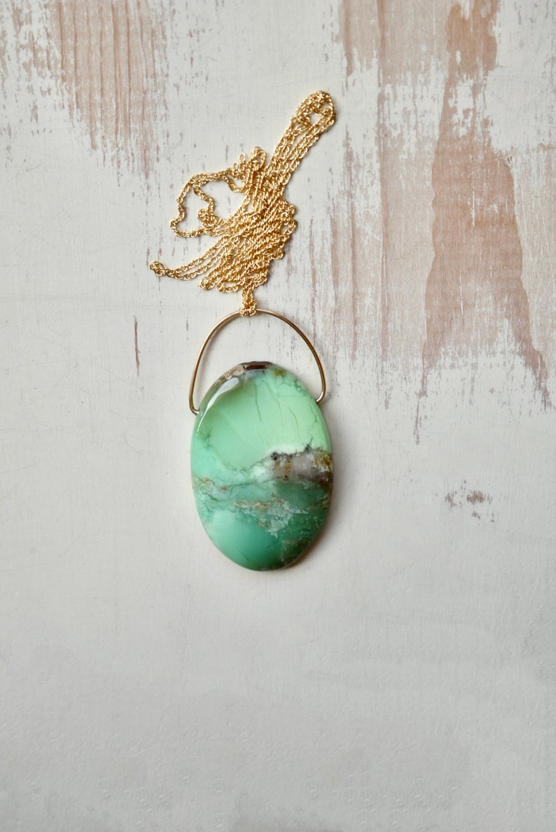 Puede incluir: Un colgante de piedra preciosa ovalado de color verde y marr&oacute;n en una cadena de oro. El colgante est&aacute; suspendido de un fino bucle de alambre de oro.