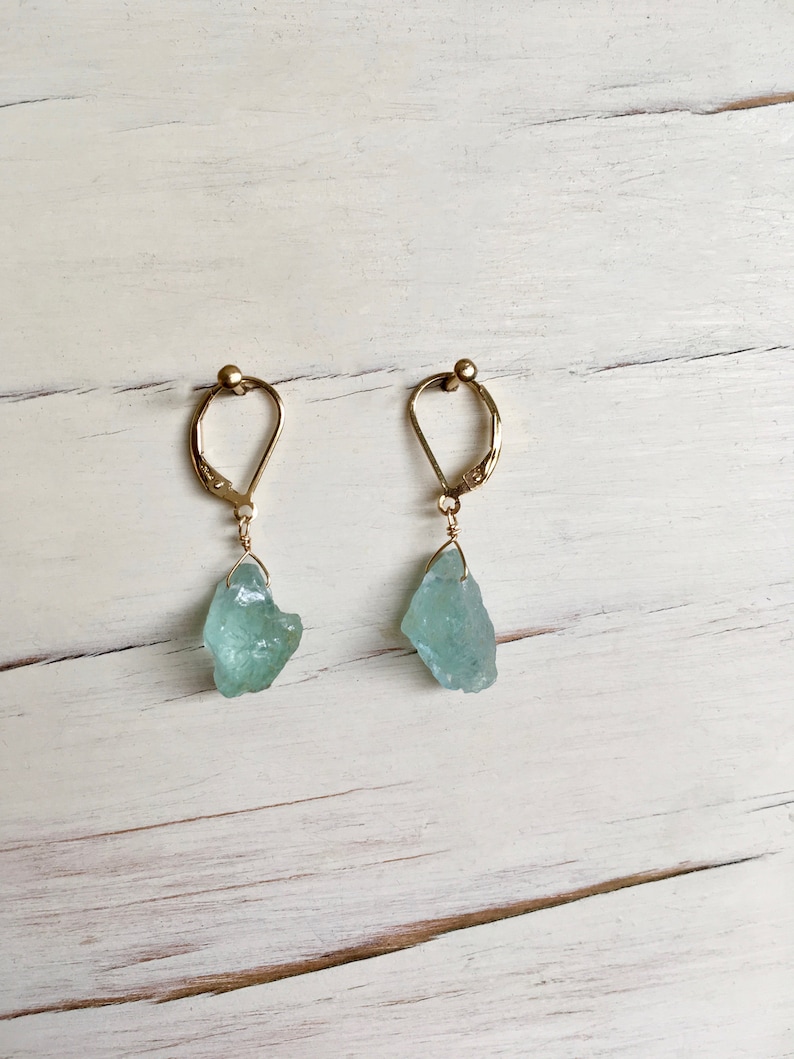 Peut inclure: Une paire de boucles d'oreilles en or avec des pierres d'aigue-marine bleu clair qui pendent. Les boucles d'oreilles sont sur un fond en bois blanc.