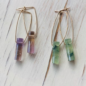 May include: A pair of gold hoop earrings with a purple and green crystal dangle. The earrings are on a white wooden surface.