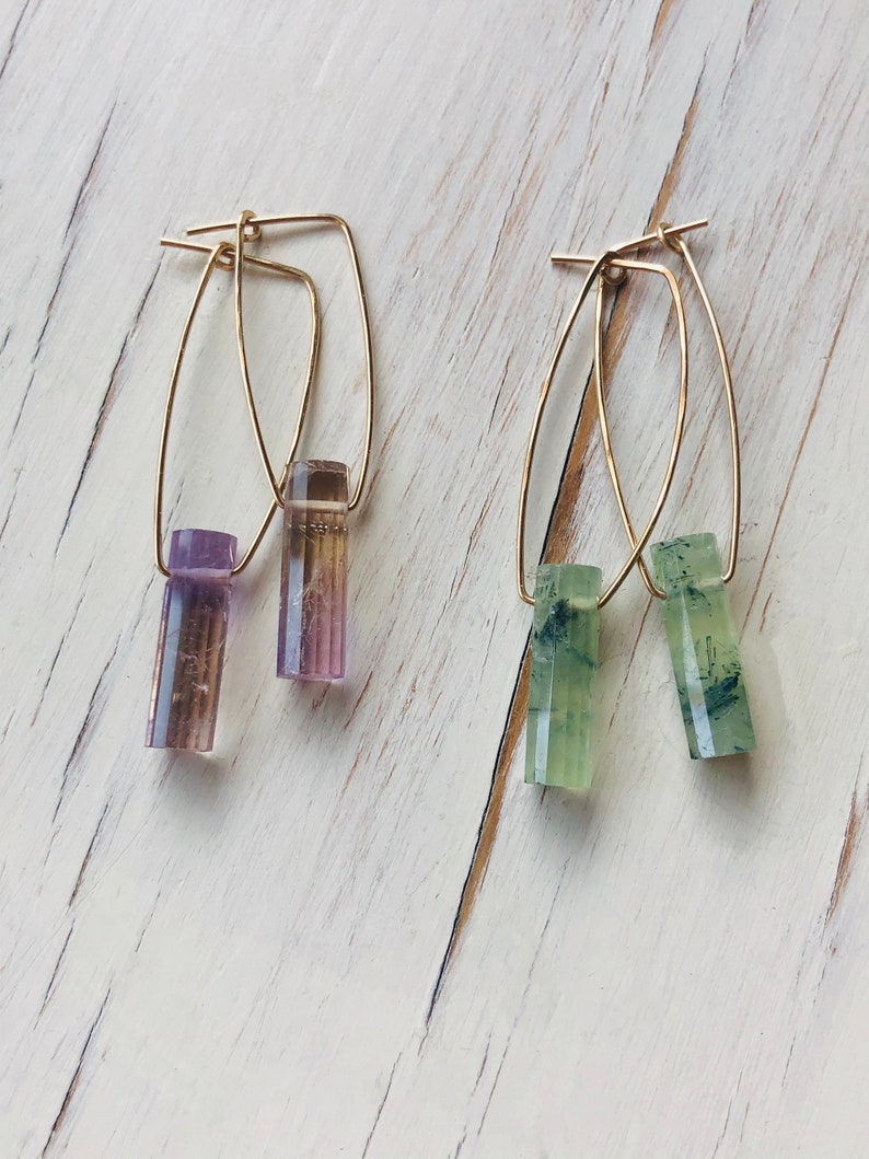May include: A pair of gold hoop earrings with a purple and green crystal dangle. The earrings are on a white wooden surface.