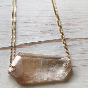 May include: A gold chain necklace with a large, clear quartz crystal pendant with brown inclusions. The crystal is faceted and has a geometric shape.