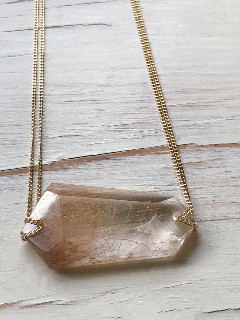 May include: A gold chain necklace with a large, clear quartz crystal pendant with brown inclusions. The crystal is faceted and has a geometric shape.