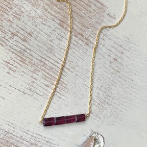 May include: A gold chain necklace with a bar pendant made of five square-shaped purple gemstones.