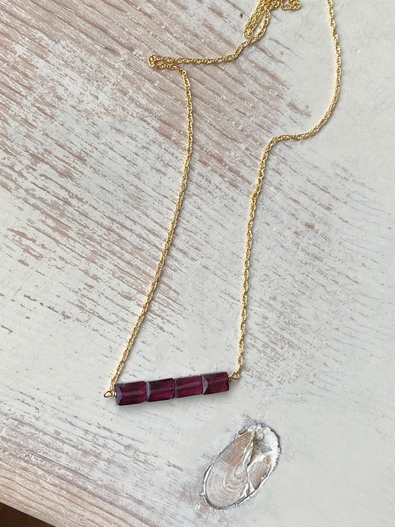 May include: A gold chain necklace with a bar pendant made of five square-shaped purple gemstones.