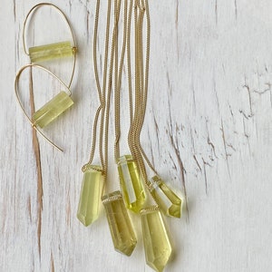 May include: Five gold necklaces with light green crystal pendants. The pendants are all different sizes and shapes. The necklaces are on a white wooden background.