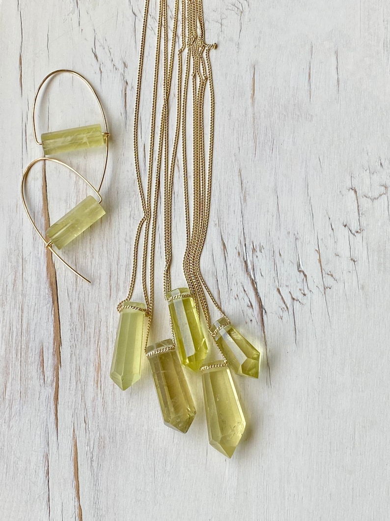 May include: Five gold necklaces with light green crystal pendants. The pendants are all different sizes and shapes. The necklaces are on a white wooden background.